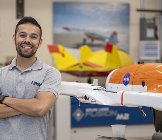 NASA Highlights: Inspiring Engineer Felipe Valdez A man wearing a gray NASA shirt posing to the left of a subscale model or an aircraft that is orange and white with the NASA meatball on it. In the background, there are aviation posters located on the wall and another subscale model on display that is yellow and red.