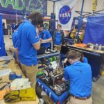 Media Invited to NASA’s Annual FIRST Robotics Competition Students from RAD Robotics Team 7111 – a FIRST Robotics team from Huntsville, Alabama, stand in blue shirts working on their robot with a large NASA meatball logo in the background.