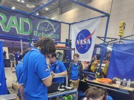 Media Invited to NASA’s Annual FIRST Robotics Competition Students from RAD Robotics Team 7111 – a FIRST Robotics team from Huntsville, Alabama, stand in blue shirts working on their robot with a large NASA meatball logo in the background.