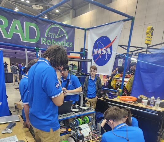 Media Invited to NASA’s Annual FIRST Robotics Competition Students from RAD Robotics Team 7111 – a FIRST Robotics team from Huntsville, Alabama, stand in blue shirts working on their robot with a large NASA meatball logo in the background.