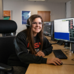 Carissa Arillo: Craft Testing and Writing NASA Manuals Carissa Arillo sits at a table with a monitor and a keyboard, wearing a headset. She smiles at the camera. On the screen is code, but it isn