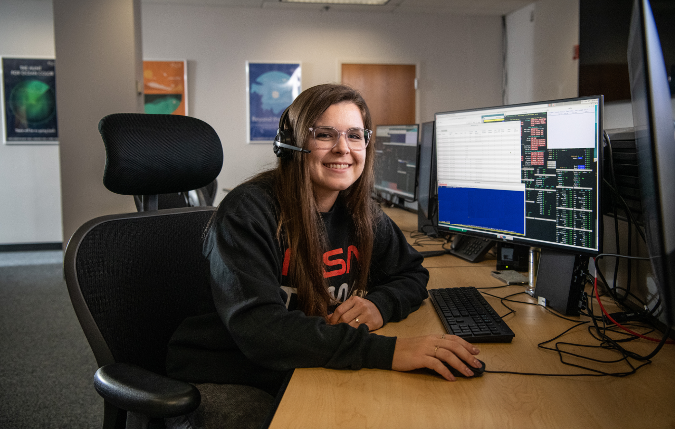 Carissa Arillo sits at a table with a monitor and a keyboard, wearing a headset. She smiles at the camera. On the screen is code, but it isn