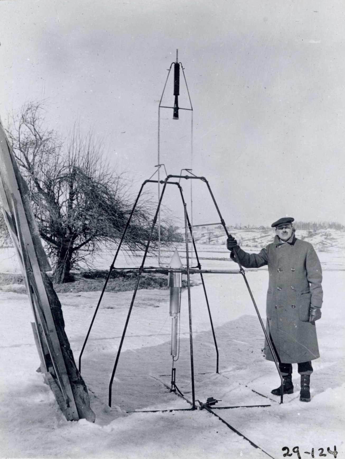 On a snowy March 16, 1926, Dr. Robert H. Goddard rests his hand on the testing frame supporting his liquid fuel rocket at Ward Farm in Auburn, Massachusetts. A wooden door is propped up at an angle next to the frame where Goddard’s assistant, Henry Sachs, later sheltered after lighting the rocket.
