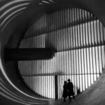 NASA’s Altitude Wind Tunnel: The Role of Turning Vanes Two men wearing hats and long coats stand at the bottom of a massive circular tunnel. At the end of the tunnel is four rows of stacked panels: this are the turning vanes. In the middle of the rows, there is a dark, cylindrical shaft that connects to the tunnel walls. This image is in black and white.