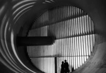 NASA’s Altitude Wind Tunnel: The Role of Turning Vanes Two men wearing hats and long coats stand at the bottom of a massive circular tunnel. At the end of the tunnel is four rows of stacked panels: this are the turning vanes. In the middle of the rows, there is a dark, cylindrical shaft that connects to the tunnel walls. This image is in black and white.
