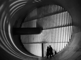 NASA’s Altitude Wind Tunnel: The Role of Turning Vanes Two men wearing hats and long coats stand at the bottom of a massive circular tunnel. At the end of the tunnel is four rows of stacked panels: this are the turning vanes. In the middle of the rows, there is a dark, cylindrical shaft that connects to the tunnel walls. This image is in black and white.