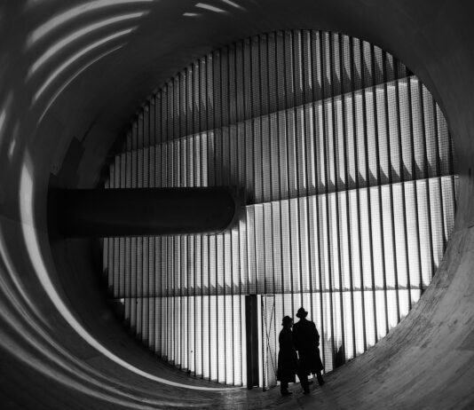 NASA’s Altitude Wind Tunnel: The Role of Turning Vanes Two men wearing hats and long coats stand at the bottom of a massive circular tunnel. At the end of the tunnel is four rows of stacked panels: this are the turning vanes. In the middle of the rows, there is a dark, cylindrical shaft that connects to the tunnel walls. This image is in black and white.