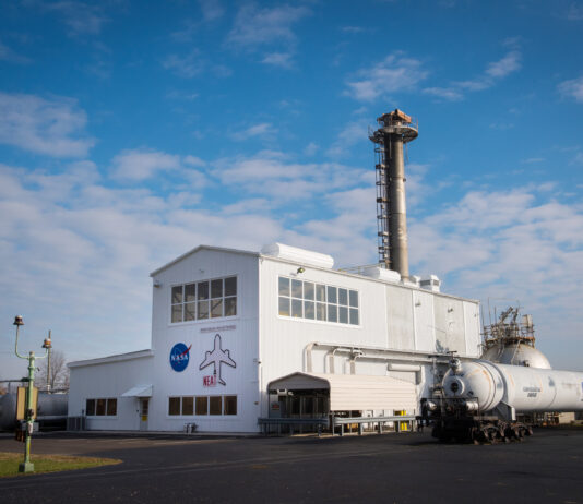 NASA Glenn Honored with Aviation Award for “NEAT” Success A metal building with an airplane and NASA logo on the front. A tall section is in the middle, a smaller flat section is on the left, and a large cylinder with tubing goes into the building on its right.