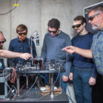 NASA Develops Its Inaugural Quantum Memory at Glenn Research Center Five engineers inspect laboratory equipment. The quantum memory the equipment holds is shown on a digital screen. Engineers wear eye protection and point at the equipment.