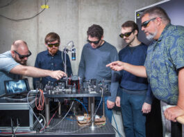 NASA Develops Its Inaugural Quantum Memory at Glenn Research Center Five engineers inspect laboratory equipment. The quantum memory the equipment holds is shown on a digital screen. Engineers wear eye protection and point at the equipment.