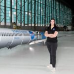 NASA Engineer Embraces Indigenous Heritage in Modern Aviation A woman wearing a black polo shirt with the NASA insignia, dark gray pants, and white shoes stands inside an aircraft hangar in front of a scaled aircraft model. The words “SUSAN Electrofan” and “PAX 180” are printed on a graphic along with fuselage, with a large NASA insignia in the center of the plane. The rear of the aircraft model, along with the wings and tail, are taken apart to display the inner components that make up its electrified propulsion system.