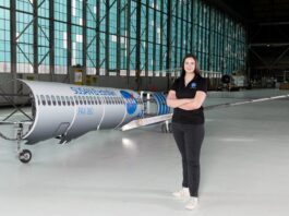 NASA Engineer Embraces Indigenous Heritage in Modern Aviation A woman wearing a black polo shirt with the NASA insignia, dark gray pants, and white shoes stands inside an aircraft hangar in front of a scaled aircraft model. The words “SUSAN Electrofan” and “PAX 180” are printed on a graphic along with fuselage, with a large NASA insignia in the center of the plane. The rear of the aircraft model, along with the wings and tail, are taken apart to display the inner components that make up its electrified propulsion system.