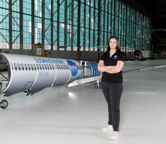 NASA Engineer Embraces Indigenous Heritage in Modern Aviation A woman wearing a black polo shirt with the NASA insignia, dark gray pants, and white shoes stands inside an aircraft hangar in front of a scaled aircraft model. The words “SUSAN Electrofan” and “PAX 180” are printed on a graphic along with fuselage, with a large NASA insignia in the center of the plane. The rear of the aircraft model, along with the wings and tail, are taken apart to display the inner components that make up its electrified propulsion system.