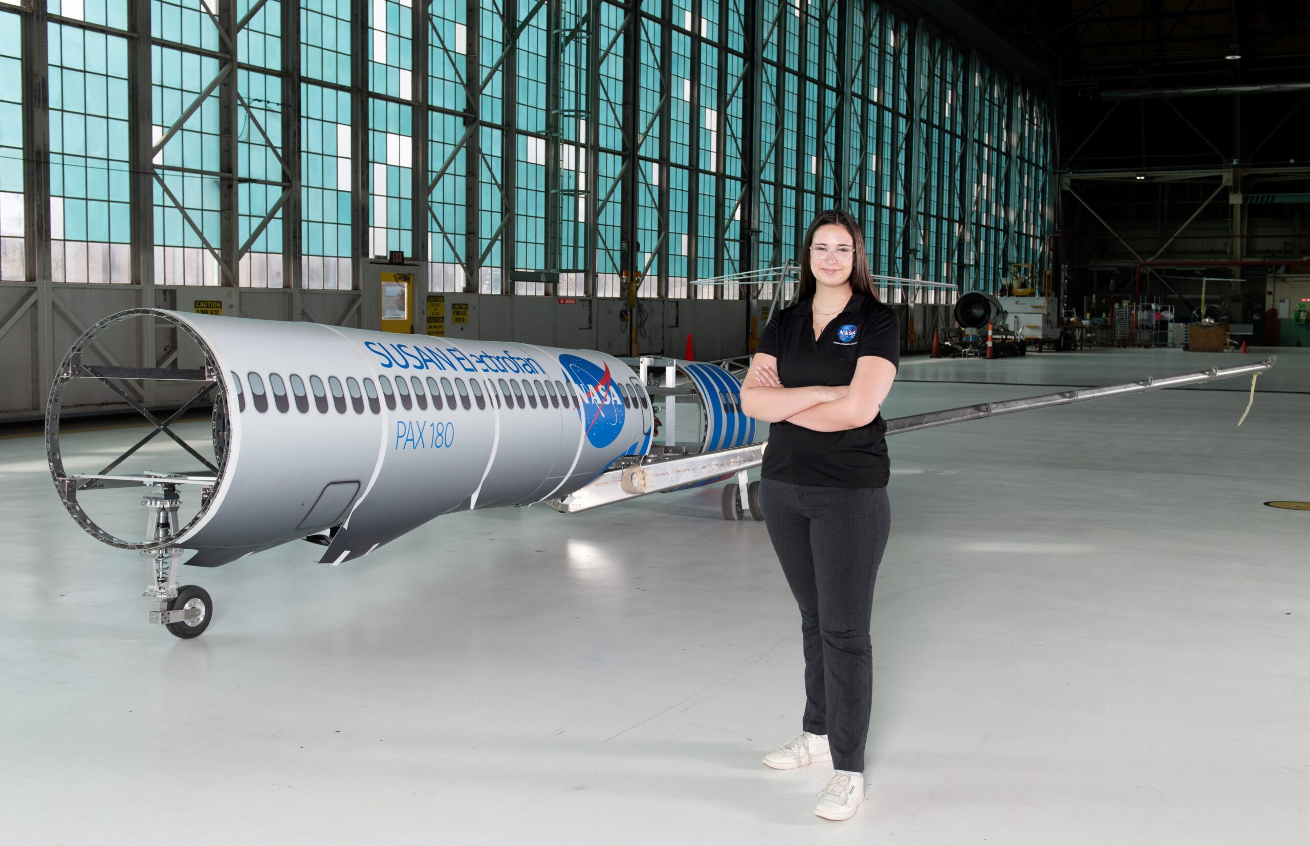 grc 2024 c 12427 002 e1731959466240.jpg A woman wearing a black polo shirt with the NASA insignia, dark gray pants, and white shoes stands inside an aircraft hangar in front of a scaled aircraft model. The words “SUSAN Electrofan” and “PAX 180” are printed on a graphic along with fuselage, with a large NASA insignia in the center of the plane. The rear of the aircraft model, along with the wings and tail, are taken apart to display the inner components that make up its electrified propulsion system.