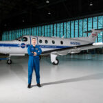 Pilot and SkillBridge Fellow Elevates NASA Research Initiatives Pilot Jeremy Johnson stands in a large hangar building in front of a blue-and-white PC-12 propeller aircraft with his arms crossed. He is wearing a dark blue NASA flight suit and black boots. Visible on the side of the plane are NASA logos, text that says, “Glenn Research Center” and “N606A,” and a small American flag.