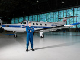 Pilot and SkillBridge Fellow Elevates NASA Research Initiatives Pilot Jeremy Johnson stands in a large hangar building in front of a blue-and-white PC-12 propeller aircraft with his arms crossed. He is wearing a dark blue NASA flight suit and black boots. Visible on the side of the plane are NASA logos, text that says, “Glenn Research Center” and “N606A,” and a small American flag.