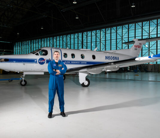 Pilot and SkillBridge Fellow Elevates NASA Research Initiatives Pilot Jeremy Johnson stands in a large hangar building in front of a blue-and-white PC-12 propeller aircraft with his arms crossed. He is wearing a dark blue NASA flight suit and black boots. Visible on the side of the plane are NASA logos, text that says, “Glenn Research Center” and “N606A,” and a small American flag.