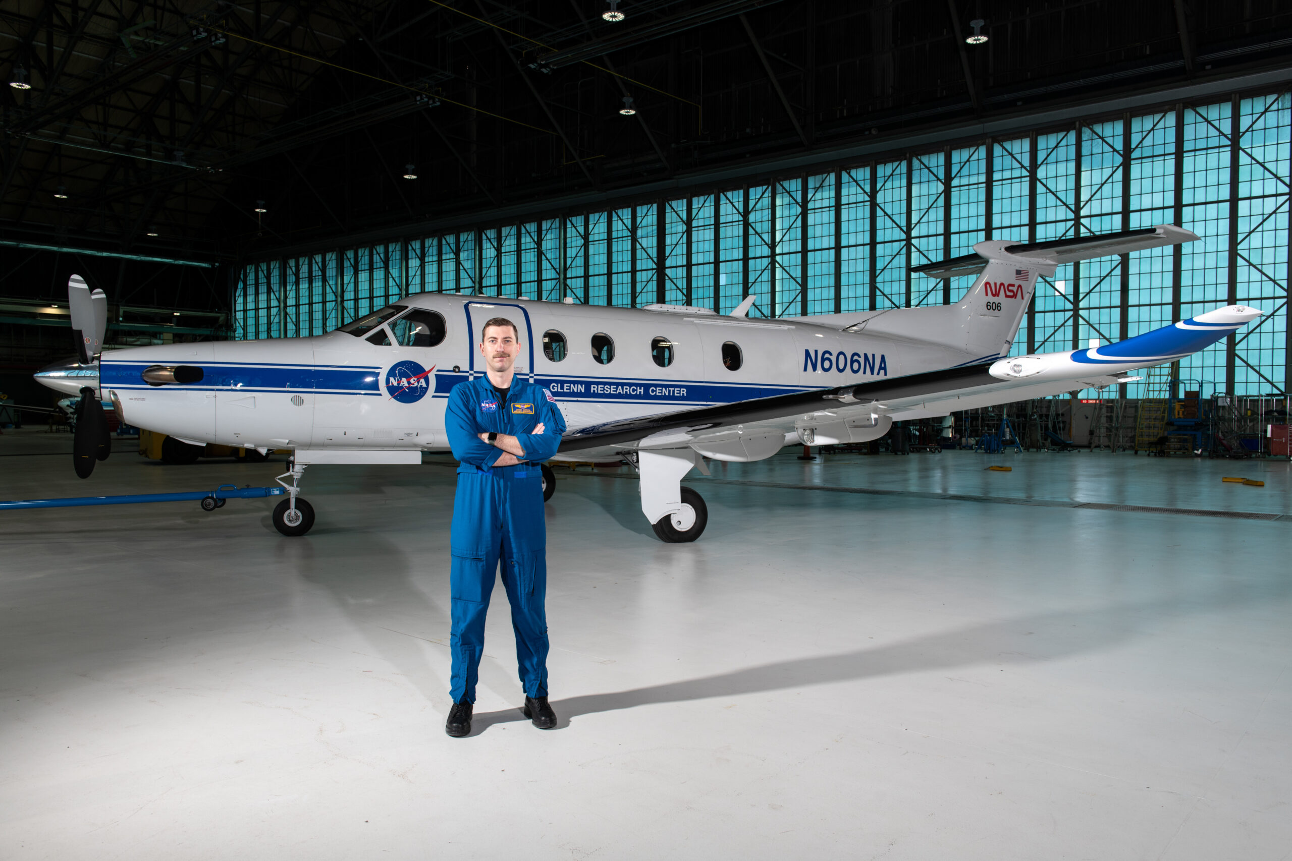 Pilot Jeremy Johnson stands in a large hangar building in front of a blue-and-white PC-12 propeller aircraft with his arms crossed. He is wearing a dark blue NASA flight suit and black boots. Visible on the side of the plane are NASA logos, text that says, “Glenn Research Center” and “N606A,” and a small American flag.