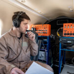 NASA Trials 5G Network for Enhanced Air Taxi Connectivity A man in a tan flight suit with a headset and microphone is sitting in a plane holding a piece of white paper. He is looking at communication equipment in the back of the plane. The equipment is comprised of large orange metal boxes with dials connected by wires to a blue computer drive rack.