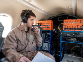 NASA Trials 5G Network for Enhanced Air Taxi Connectivity A man in a tan flight suit with a headset and microphone is sitting in a plane holding a piece of white paper. He is looking at communication equipment in the back of the plane. The equipment is comprised of large orange metal boxes with dials connected by wires to a blue computer drive rack.