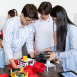 Apply for NASA Glenn’s Free Summer Engineering Institute now! Three High School Engineering Institute students are centered in the foreground of the image working on a small vehicle with large red plastic wheels. There are two more students in the background working on their own identical vehicle.