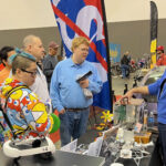NASA Glenn Unveils Stirling Engine at Auto-Rama Event A NASA subject matter expert stands behind a display table and explains the technology behind free-standing piston Stirling engines to a group of people.