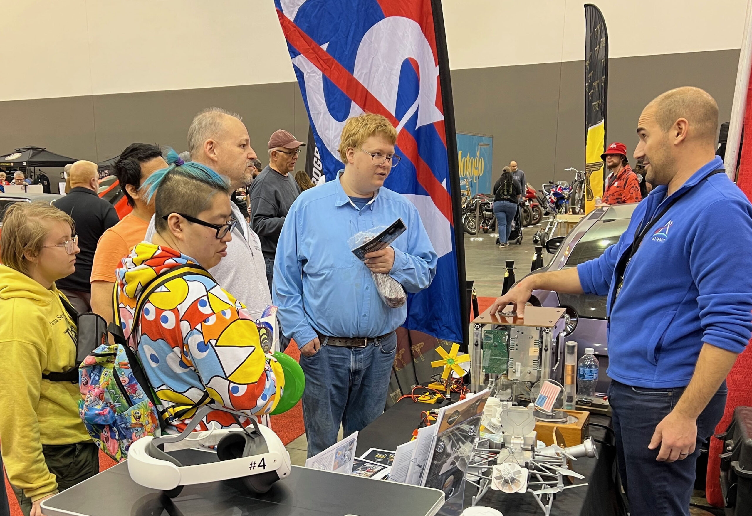 grc 2025 cn 00012.jpg A NASA subject matter expert stands behind a display table and explains the technology behind free-standing piston Stirling engines to a group of people.