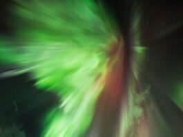 AI Collaborations with NASA Enhance Solar Event Predictions The green lights of an aurora dramatically explode outward against the backdrop of the night sky peppered with fluffy white clouds and pinprick stars. A hint of red is also visible in the center of the light. Pine trees cast in shadow are seen below.