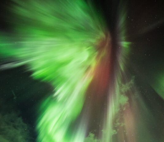 AI Collaborations with NASA Enhance Solar Event Predictions The green lights of an aurora dramatically explode outward against the backdrop of the night sky peppered with fluffy white clouds and pinprick stars. A hint of red is also visible in the center of the light. Pine trees cast in shadow are seen below.