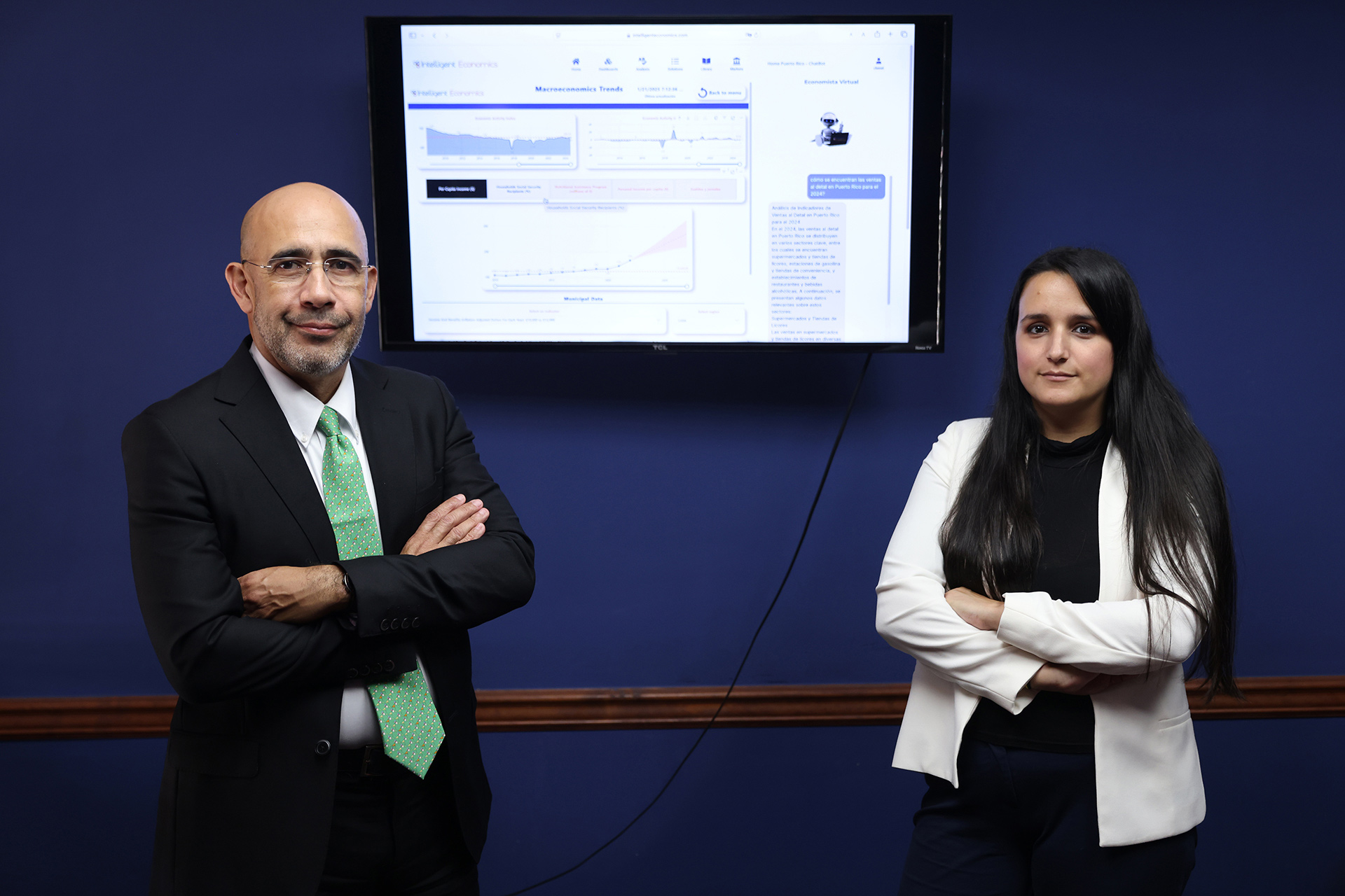 gustavo velez chantal benet 2.jpg A man and a woman stand in front of a blue wall with a computer monitor between them