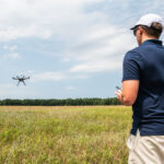 NASA Advances Drone Safety with New Management System In the right foreground of the image a male wears a navy blue polo shirt and tan shorts and he faces away from the viewer. His left side is in view and he is wearing a white hat and black sunglasses. He holds a white controller. In the background is a field and a far away tree line. A small, black drone flies to the left side of the image with blue sky and white clouds behind the drone.