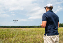 NASA Advances Drone Safety with New Management System In the right foreground of the image a male wears a navy blue polo shirt and tan shorts and he faces away from the viewer. His left side is in view and he is wearing a white hat and black sunglasses. He holds a white controller. In the background is a field and a far away tree line. A small, black drone flies to the left side of the image with blue sky and white clouds behind the drone.