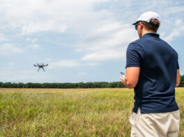 NASA Advances Drone Safety with New Management System In the right foreground of the image a male wears a navy blue polo shirt and tan shorts and he faces away from the viewer. His left side is in view and he is wearing a white hat and black sunglasses. He holds a white controller. In the background is a field and a far away tree line. A small, black drone flies to the left side of the image with blue sky and white clouds behind the drone.