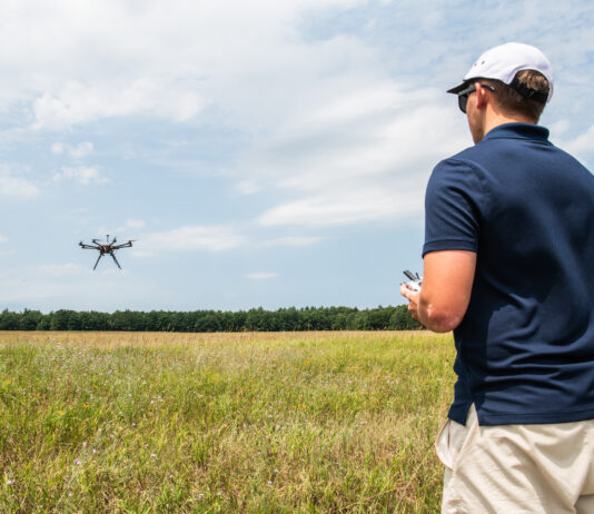 NASA Advances Drone Safety with New Management System In the right foreground of the image a male wears a navy blue polo shirt and tan shorts and he faces away from the viewer. His left side is in view and he is wearing a white hat and black sunglasses. He holds a white controller. In the background is a field and a far away tree line. A small, black drone flies to the left side of the image with blue sky and white clouds behind the drone.