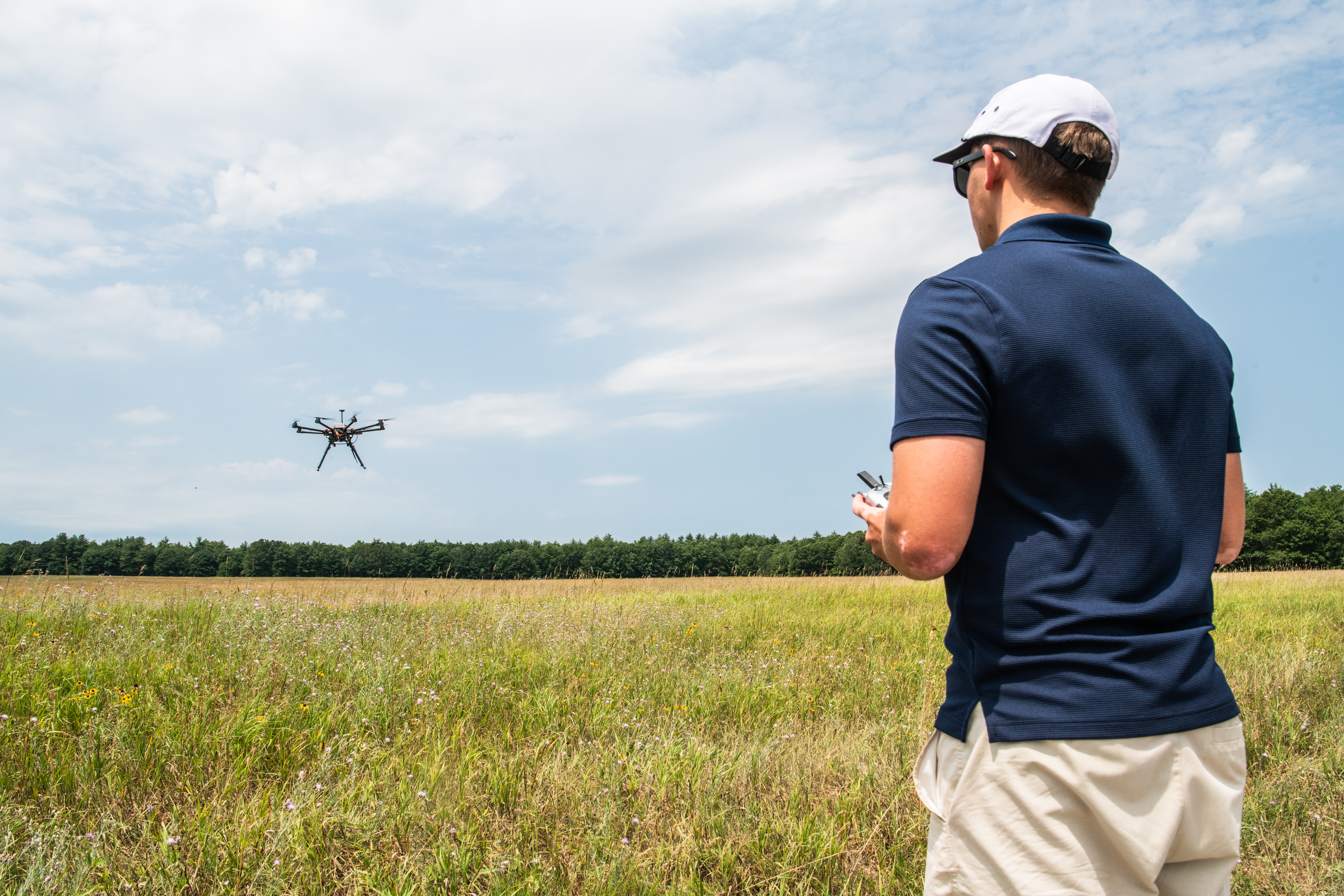 In the right foreground of the image a male wears a navy blue polo shirt and tan shorts and he faces away from the viewer. His left side is in view and he is wearing a white hat and black sunglasses. He holds a white controller. In the background is a field and a far away tree line. A small, black drone flies to the left side of the image with blue sky and white clouds behind the drone.