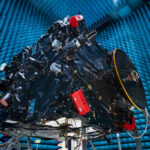 AI-Powered Hera Communicates Earth Defense Against Asteroids A large rectangular structure with many attachments and complex parts suspended in an enormous hangar; a woman in a sterile robe and hair cover stands on the ground at lower right.