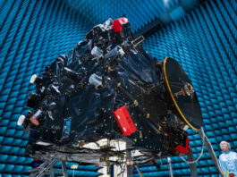 AI-Powered Hera Communicates Earth Defense Against Asteroids A large rectangular structure with many attachments and complex parts suspended in an enormous hangar; a woman in a sterile robe and hair cover stands on the ground at lower right.