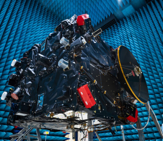 AI-Powered Hera Communicates Earth Defense Against Asteroids A large rectangular structure with many attachments and complex parts suspended in an enormous hangar; a woman in a sterile robe and hair cover stands on the ground at lower right.