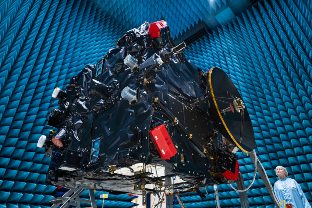 hera msft 1 1024x683.jpg A large rectangular structure with many attachments and complex parts suspended in an enormous hangar; a woman in a sterile robe and hair cover stands on the ground at lower right.