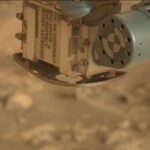 Exploring the Canyon: Sols 4471-4472 NASA Science Update A color close-up photo from the Martian surface shows a blurred-out background of rocky terrain colored pale orange-tan, while the top center and right corner of the image are filled with a view of a rover instrument in close focus. A metallic sage green cylinder extends from the upper right corner toward the center of the frame; its circular face has a line of nine holes in the metal around its outer edge, and inside that is a target formed by two concentric circles, each of them divided in quarters of alternating green and white. The cylinder is attached to the bulk of the instrument, which is sort of a rounded cube shape, creamy tan in color and dusted in some spots with brown-orange soil. This main part of the instrument dominates the upper middle half of the image, with one edge facing the viewer. The right-side face of the cube shows four small hex screws staggered up one side, and a pair of delicate wires, braided and leading to the green cylinder. The left face of the cube shows several lines of handwritten numbers and capital letters in dark gray ink or paint, rotated 90 degrees to the right from the viewer’s perspective. Part of the inscription reads “APXS SENSOR HEAD ASSEMBLY.”