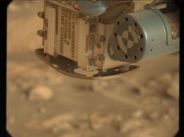 Exploring the Canyon: Sols 4471-4472 NASA Science Update A color close-up photo from the Martian surface shows a blurred-out background of rocky terrain colored pale orange-tan, while the top center and right corner of the image are filled with a view of a rover instrument in close focus. A metallic sage green cylinder extends from the upper right corner toward the center of the frame; its circular face has a line of nine holes in the metal around its outer edge, and inside that is a target formed by two concentric circles, each of them divided in quarters of alternating green and white. The cylinder is attached to the bulk of the instrument, which is sort of a rounded cube shape, creamy tan in color and dusted in some spots with brown-orange soil. This main part of the instrument dominates the upper middle half of the image, with one edge facing the viewer. The right-side face of the cube shows four small hex screws staggered up one side, and a pair of delicate wires, braided and leading to the green cylinder. The left face of the cube shows several lines of handwritten numbers and capital letters in dark gray ink or paint, rotated 90 degrees to the right from the viewer’s perspective. Part of the inscription reads “APXS SENSOR HEAD ASSEMBLY.”