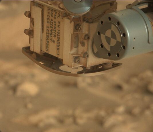Exploring the Canyon: Sols 4471-4472 NASA Science Update A color close-up photo from the Martian surface shows a blurred-out background of rocky terrain colored pale orange-tan, while the top center and right corner of the image are filled with a view of a rover instrument in close focus. A metallic sage green cylinder extends from the upper right corner toward the center of the frame; its circular face has a line of nine holes in the metal around its outer edge, and inside that is a target formed by two concentric circles, each of them divided in quarters of alternating green and white. The cylinder is attached to the bulk of the instrument, which is sort of a rounded cube shape, creamy tan in color and dusted in some spots with brown-orange soil. This main part of the instrument dominates the upper middle half of the image, with one edge facing the viewer. The right-side face of the cube shows four small hex screws staggered up one side, and a pair of delicate wires, braided and leading to the green cylinder. The left face of the cube shows several lines of handwritten numbers and capital letters in dark gray ink or paint, rotated 90 degrees to the right from the viewer’s perspective. Part of the inscription reads “APXS SENSOR HEAD ASSEMBLY.”
