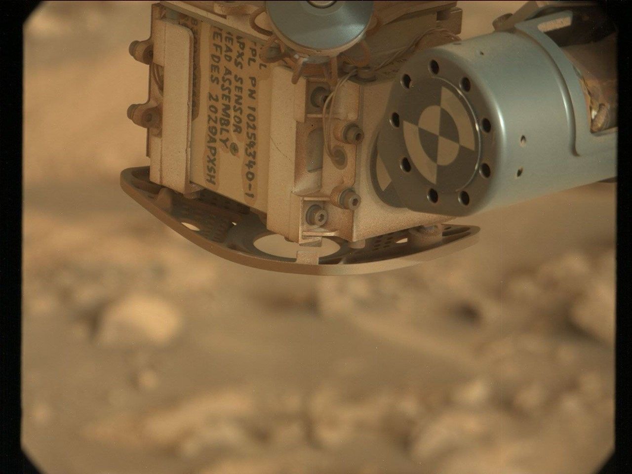 A color close-up photo from the Martian surface shows a blurred-out background of rocky terrain colored pale orange-tan, while the top center and right corner of the image are filled with a view of a rover instrument in close focus. A metallic sage green cylinder extends from the upper right corner toward the center of the frame; its circular face has a line of nine holes in the metal around its outer edge, and inside that is a target formed by two concentric circles, each of them divided in quarters of alternating green and white. The cylinder is attached to the bulk of the instrument, which is sort of a rounded cube shape, creamy tan in color and dusted in some spots with brown-orange soil. This main part of the instrument dominates the upper middle half of the image, with one edge facing the viewer. The right-side face of the cube shows four small hex screws staggered up one side, and a pair of delicate wires, braided and leading to the green cylinder. The left face of the cube shows several lines of handwritten numbers and capital letters in dark gray ink or paint, rotated 90 degrees to the right from the viewer’s perspective. Part of the inscription reads “APXS SENSOR HEAD ASSEMBLY.”