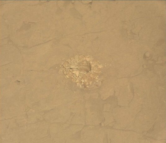 Examining Altadena Drill Site: NASA’s Curiosity Insights A color close-up photo of the Martian surface shows flat, mostly smooth, tan-orange ground, crisscrossed with a few shallow grooves and indentations. At the center is a hole cored into the ground, surrounded by a pile of soil and small rocks dug from the hole — those are similarly colored to the surrounding ground, but in shades ranging from lighter and more yellow to darker and deeper orange. Some of the back wall of the interior of the hole is also visible.