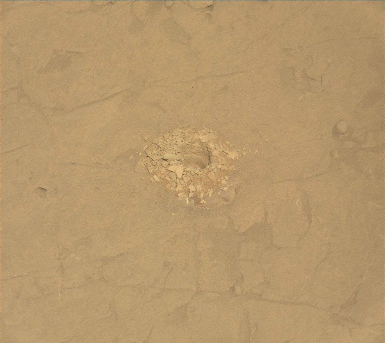 https mars.nasa .gov msl raw images msss 04564 mcam 4564mr1077420012302300e01 dxxx.jpg A color close-up photo of the Martian surface shows flat, mostly smooth, tan-orange ground, crisscrossed with a few shallow grooves and indentations. At the center is a hole cored into the ground, surrounded by a pile of soil and small rocks dug from the hole — those are similarly colored to the surrounding ground, but in shades ranging from lighter and more yellow to darker and deeper orange. Some of the back wall of the interior of the hole is also visible.