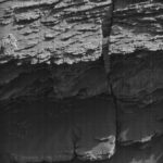 NASA: Volcán Peña Blanca’s Complex Structure Unveiled on Mars A grayscale photo from the Martian surface shows a rocky outcrop rising from sandy ground, all of it medium gray. The rock feature is composed of thin, rough-edged layers, stacked on top of each other, rising from the ground to the top of the frame. The layer at bottom sticks out farther, toward the viewer, and is thicker than the rest and covered in a layer of sand.