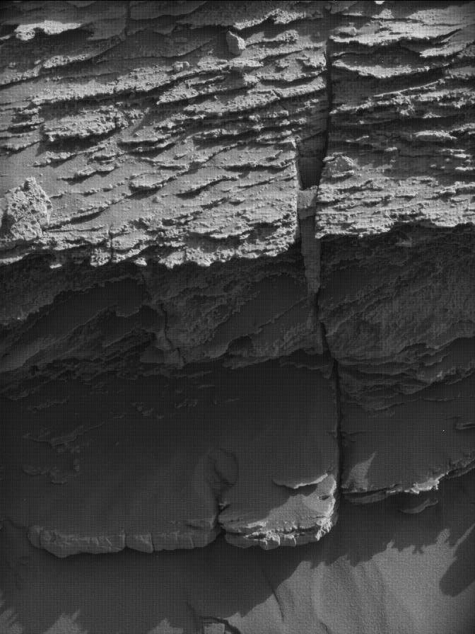 https mars.nasa .gov msl raw images msss 04591 mcam 4591ml1079010041802929c00 dxxx.jpg A grayscale photo from the Martian surface shows a rocky outcrop rising from sandy ground, all of it medium gray. The rock feature is composed of thin, rough-edged layers, stacked on top of each other, rising from the ground to the top of the frame. The layer at bottom sticks out farther, toward the viewer, and is thicker than the rest and covered in a layer of sand.