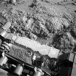 Sol 4553: Revisiting the Boxwork Formation – NASA Update A grayscale photograph of Martian terrain in front of the Curiosity rover shows very bumpy, uneven ground with large, polygonal sections of rock on the surface separated by gouges in the soil. The bumpy area and a smoother patch just beyond that all appear shiny. The bottom of the frame shows parts of the rover, running from the middle left edge to the lower right corner of the image, including part of its robotic arm which carries a nameplate imprinted with “Curiosity” outlined in white, all-capital letters, and to the right of that a line drawing of the rover.