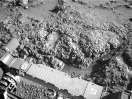 Sol 4553: Revisiting the Boxwork Formation – NASA Update A grayscale photograph of Martian terrain in front of the Curiosity rover shows very bumpy, uneven ground with large, polygonal sections of rock on the surface separated by gouges in the soil. The bumpy area and a smoother patch just beyond that all appear shiny. The bottom of the frame shows parts of the rover, running from the middle left edge to the lower right corner of the image, including part of its robotic arm which carries a nameplate imprinted with “Curiosity” outlined in white, all-capital letters, and to the right of that a line drawing of the rover.