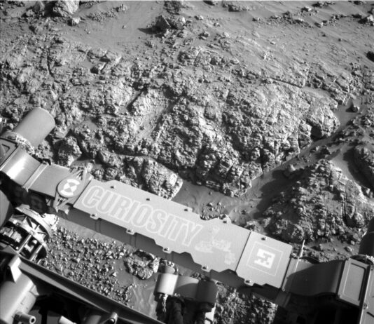 Sol 4553: Revisiting the Boxwork Formation – NASA Update A grayscale photograph of Martian terrain in front of the Curiosity rover shows very bumpy, uneven ground with large, polygonal sections of rock on the surface separated by gouges in the soil. The bumpy area and a smoother patch just beyond that all appear shiny. The bottom of the frame shows parts of the rover, running from the middle left edge to the lower right corner of the image, including part of its robotic arm which carries a nameplate imprinted with “Curiosity” outlined in white, all-capital letters, and to the right of that a line drawing of the rover.